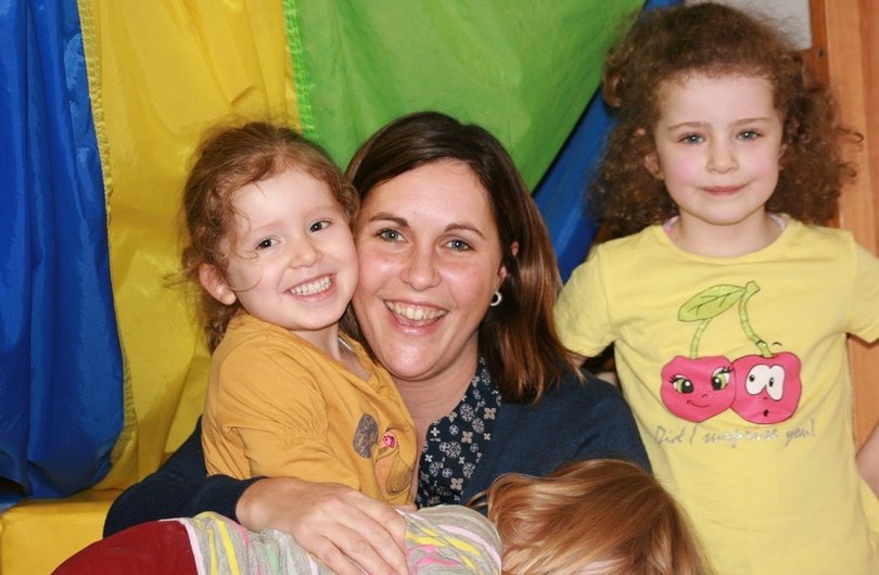 Auf dem Bild sind eine Kinderpflegerin und drei kleine Kinder zu sehen. Die Kinderpflegerin sowie zwei der Kinder lächeln in die Kamera, während das dritte Kind auf den Beinen der Pflegerin liegt und zu ihr schaut.