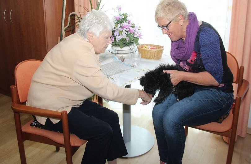 Auf dem Bild sind eine ältere Dame und eine Betreuungskraft in einem Pflegeheim zu sehen. Beide sitzen auf einem Stuhl, im Hintergrund ist ein Kaffeetisch zu sehen, auf welchem eine aufgeschlagene Fernsehzeitung liegt. Die Betreuungskraft hält einen schwarzen Hund auf ihrem Schoß, welcher an der Hand der älteren Dame schnüffelt.