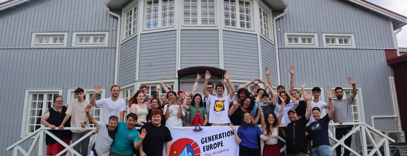 Zu sehen ist ein Bild, welches beim Treffen in Finnland aufgenommen wurde. Auf dem Bild sind alle Teilnehmer vor dem gemeinsam bewohnten Haus zu sehen. In der Mitte wird die Flagge des Projekts "Generation Europe" hochgehalten.